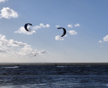 Kolorowe latawce nad zatoką pokazują doskonałe warunki do sport&oacute;w wodnych w Jastarni. Dynamiczny kadr prezentuje kitesurfing na P&oacute;łwyspie Helskim w wietrzny dzień.