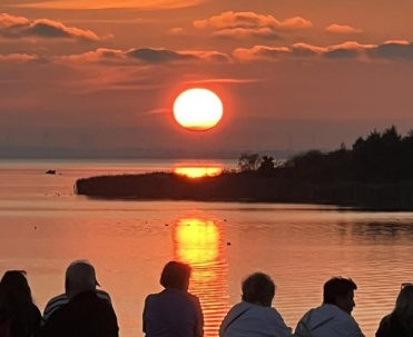 Sylwetki os&oacute;b stojących nad wodą podkreślają skalę spektakularnego zachodu słońca. Zdjęcie pokazuje, dlaczego wieczory nad Zatoką Pucką w Jastarni przyciągają spacerowicz&oacute;w.